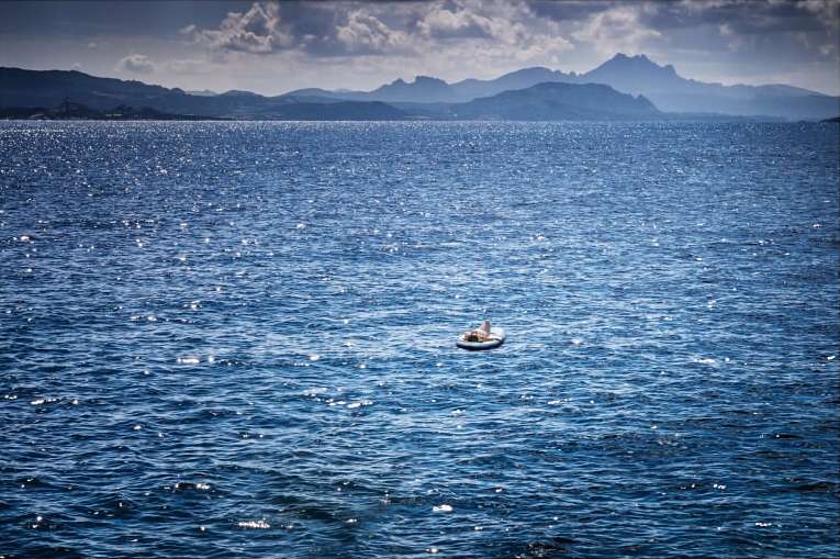 Vast blue ocean glistens with sun, person rests on an inflatable raft, mountains in the distance under cloudy sky, relaxation scene.