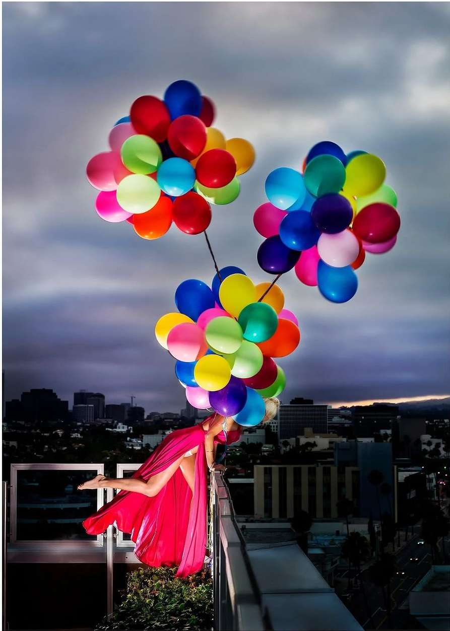 An adult female in a pink dress stands on a rooftop with a cityscape backdrop, holding a large collection of colorful balloons.