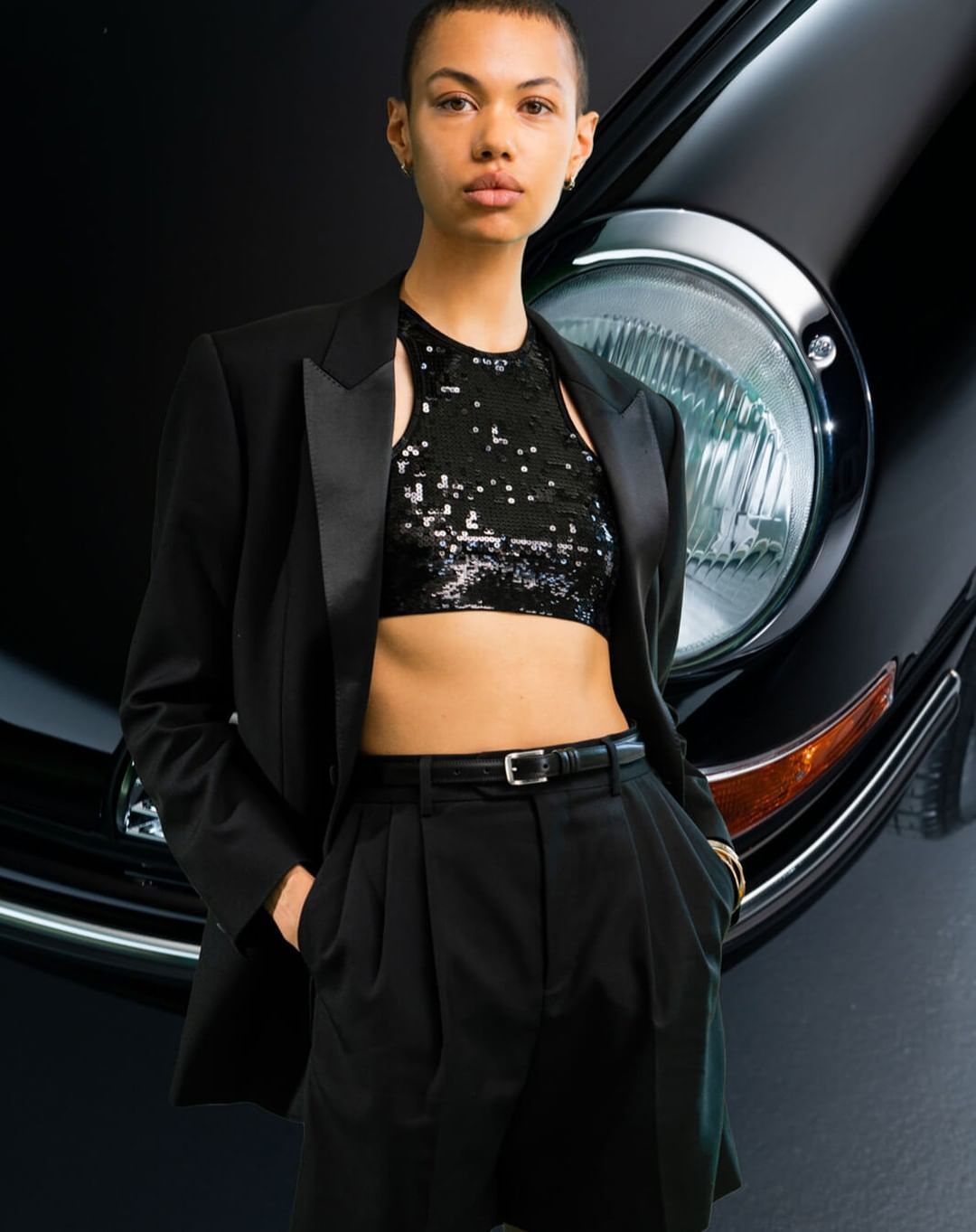 A woman in a black blazer and sequin crop top stands in front of a black car with a headlight in an indoor studio setting.