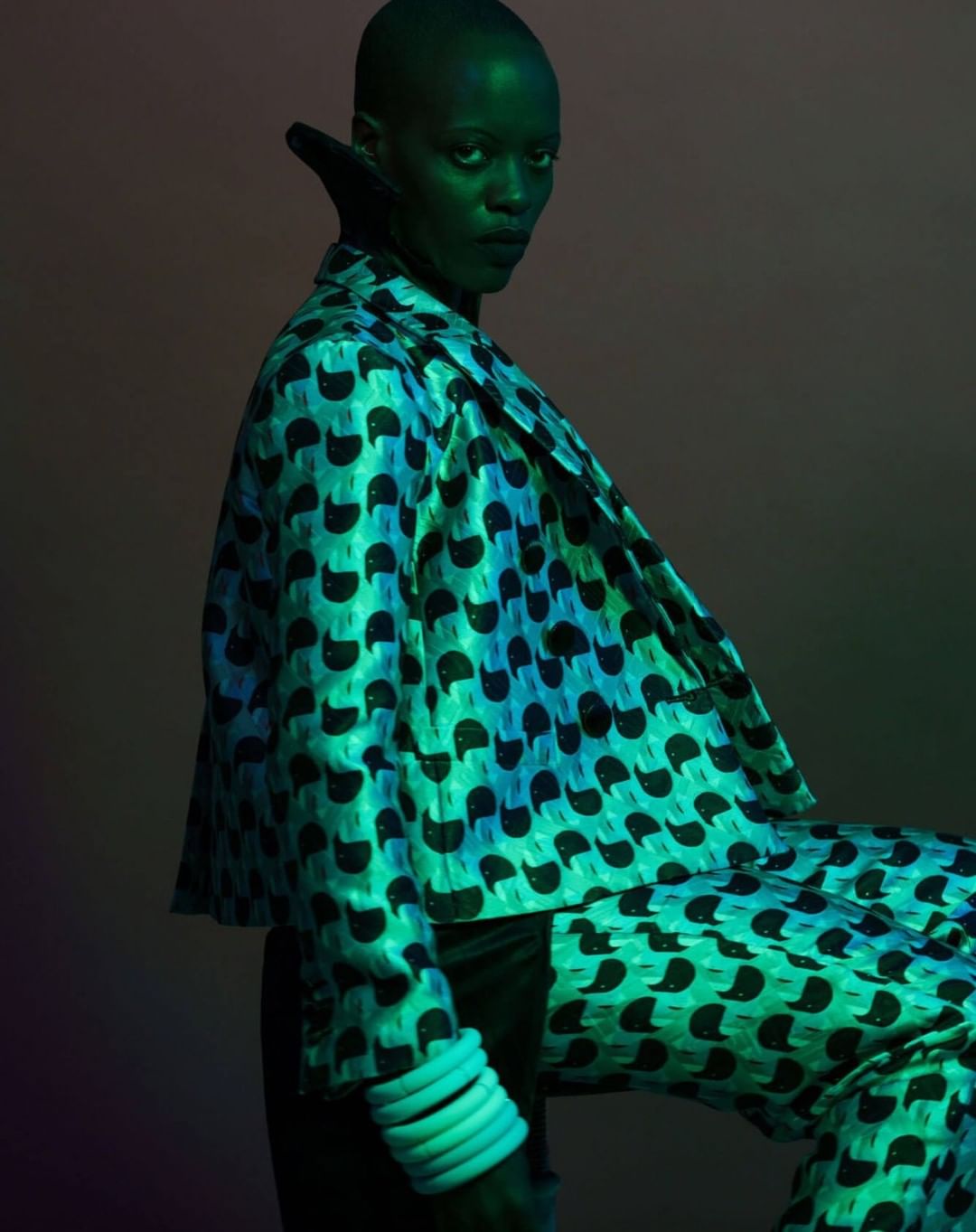 Studio shot of a stylish woman in a patterned suit with green and blue lighting, accessorized with bangles and a high collar.