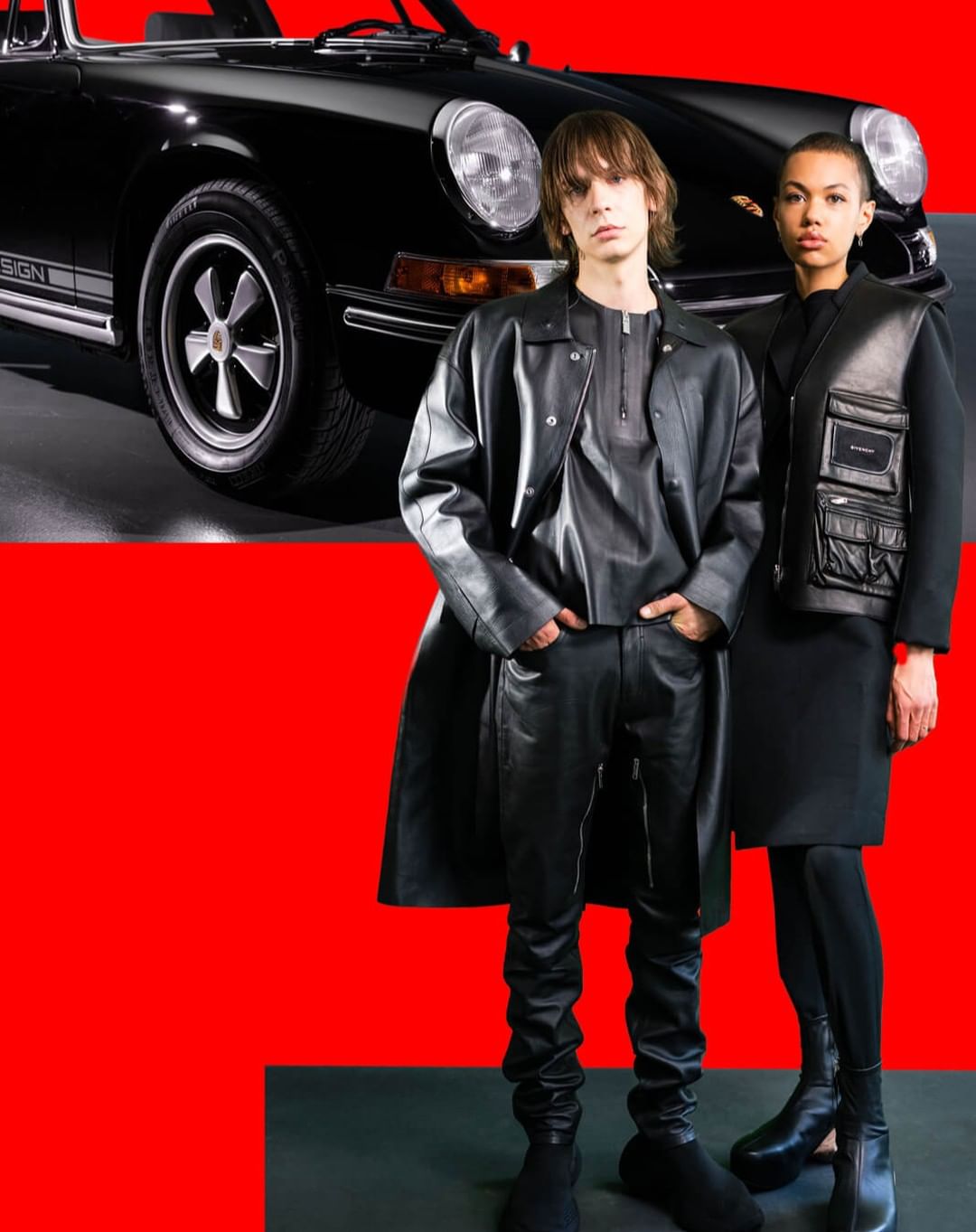 Editorial fashion shot with a male model in a black leather outfit, a female model, and a black Porsche against a red backdrop.