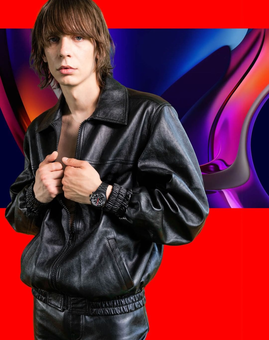 An adult male poses in a black leather jacket and pants against a background of red and colorful, abstract shapes. He is wearing a watch.