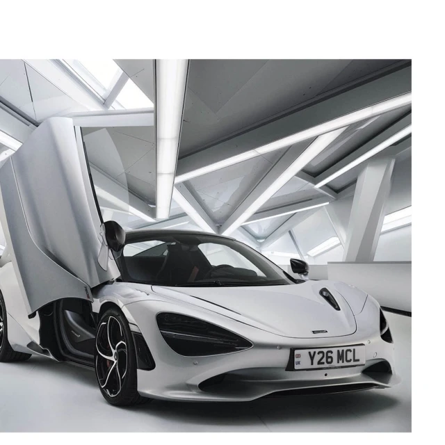 Silver Mclaren sports car with open doors, displayed in an indoor showroom with geometric architectural background lighting.