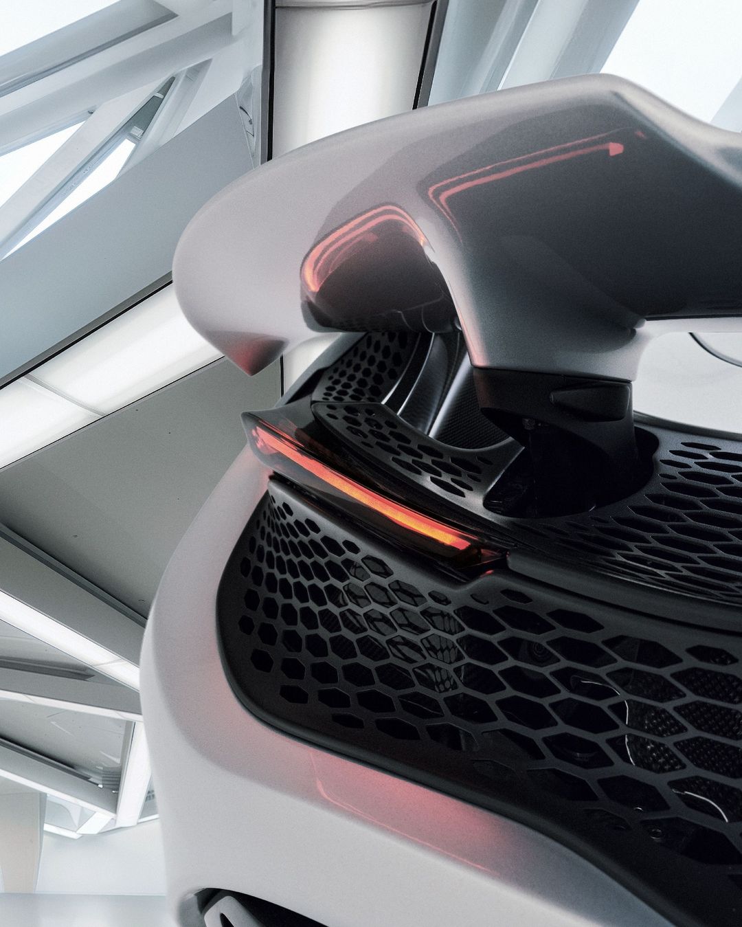 Close-up of a futuristic white car's rear with its spoiler, black hexagonal patterned grill, and illuminated brake lights, architectural setting.