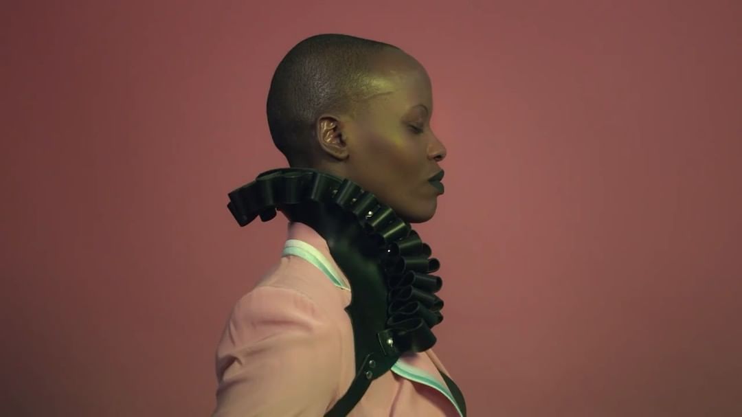 Close-up profile shot of a Black adult female with a shaved head wearing a ruffled black leather harness over a pink jacket on a coral background.