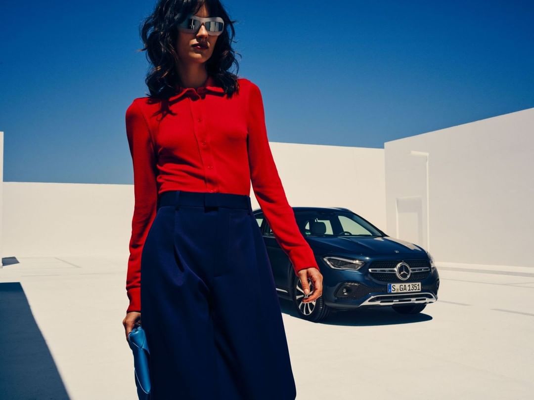 Stylish woman in red blouse and navy skirt poses with a dark blue Mercedes-Benz against a bright blue sky and white architectural backdrop.