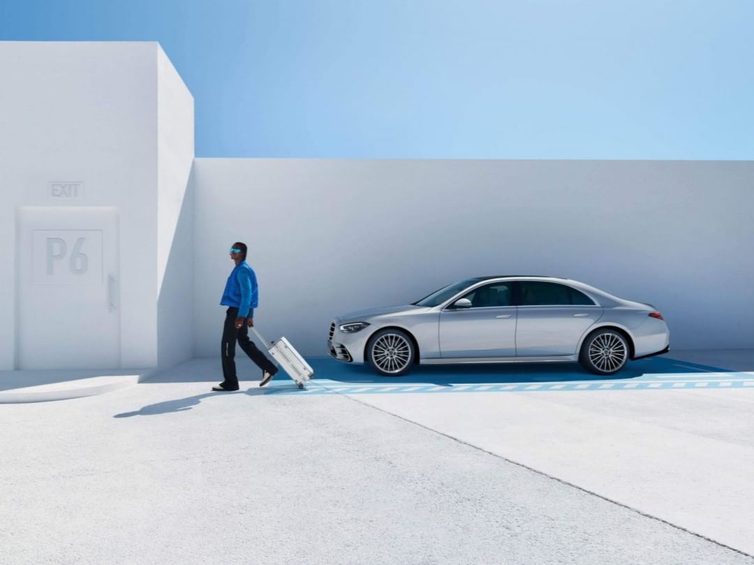 An adult man pulls rolling luggage near a silver sedan in front of a white parking garage against a light blue sky on a bright day.