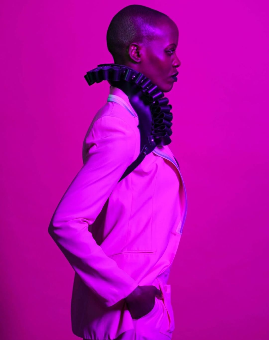 Striking profile of a bald Black woman in a pink suit and ruffled collar against a vibrant pink backdrop, showcasing a fashion-forward aesthetic.