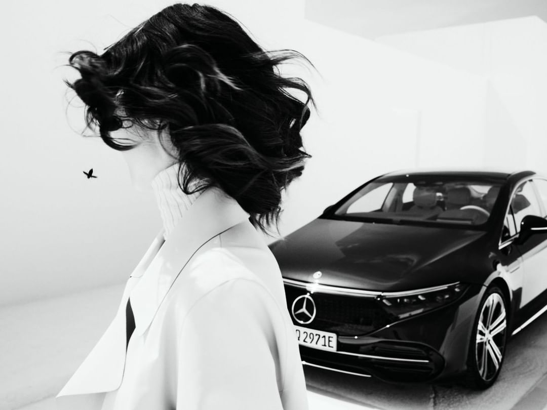 Black and white photo of a woman with dark hair standing next to a black Mercedes-Benz EQS, showcasing the car's design.