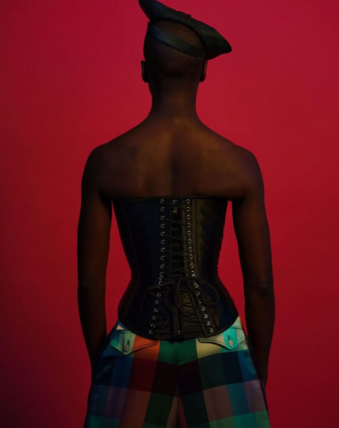 A person wearing a black corset and plaid pants stands against a vibrant red backdrop, showcasing fashion and style. Back view.