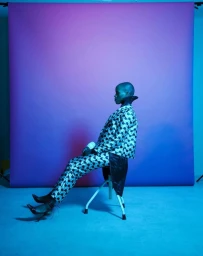 A bald Black woman in a patterned jacket and matching trousers seated on a white stool against a purple gradient backdrop.