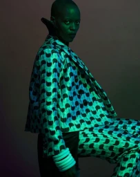 Portrait of a bald person wearing a patterned turquoise blazer and matching trousers under green lighting