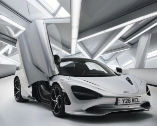 Silver sports car with upward-opening butterfly doors parked inside a modern minimalist showroom with angled lighting fixtures.