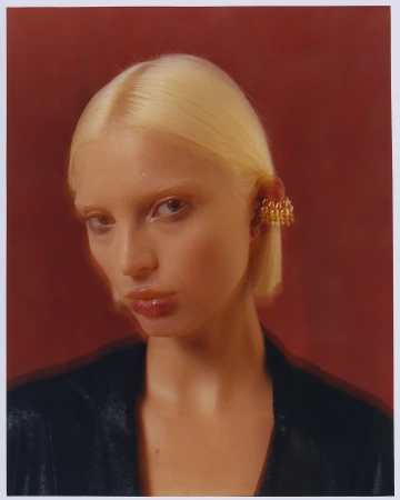 Close-up portrait of a fair-skinned blonde woman with sleek hair, wearing an ornate gold ear cuff and a black jacket against a red backdrop.
