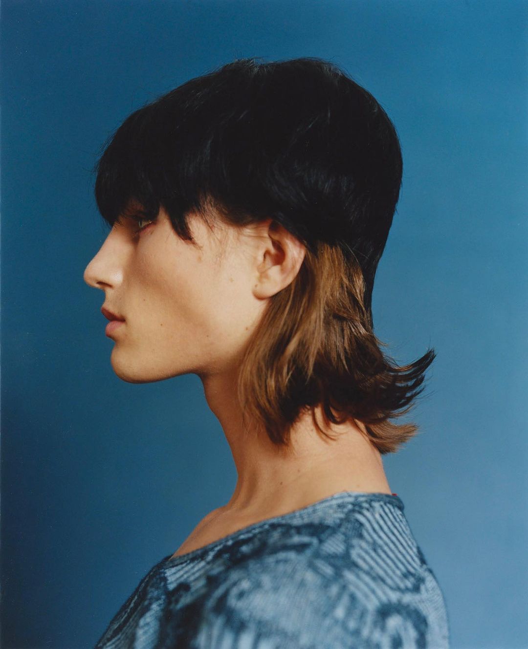 Side portrait of an adult with short dark hair and blue patterned top against a blue background in a fashion photography style image.