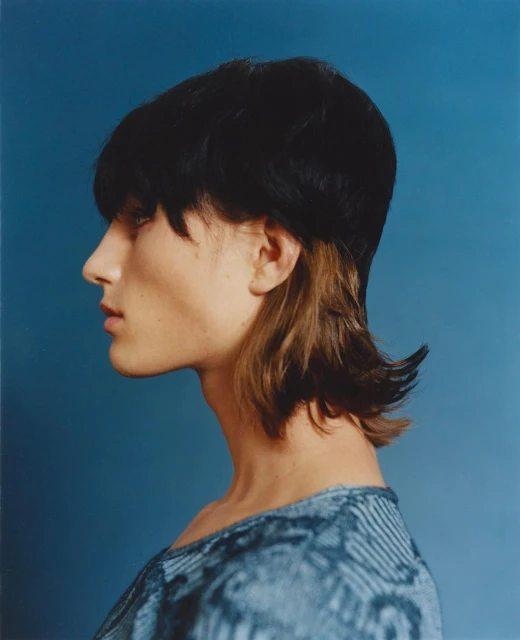 Side portrait of an adult with short dark hair and blue patterned top against a blue background in a fashion photography style image.