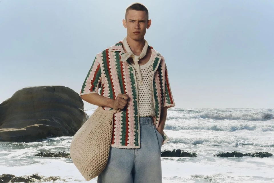 A fair-skinned adult male model poses on a rocky beach, wearing a colorful crochet shirt, light-wash denim pants, and a woven tote bag.