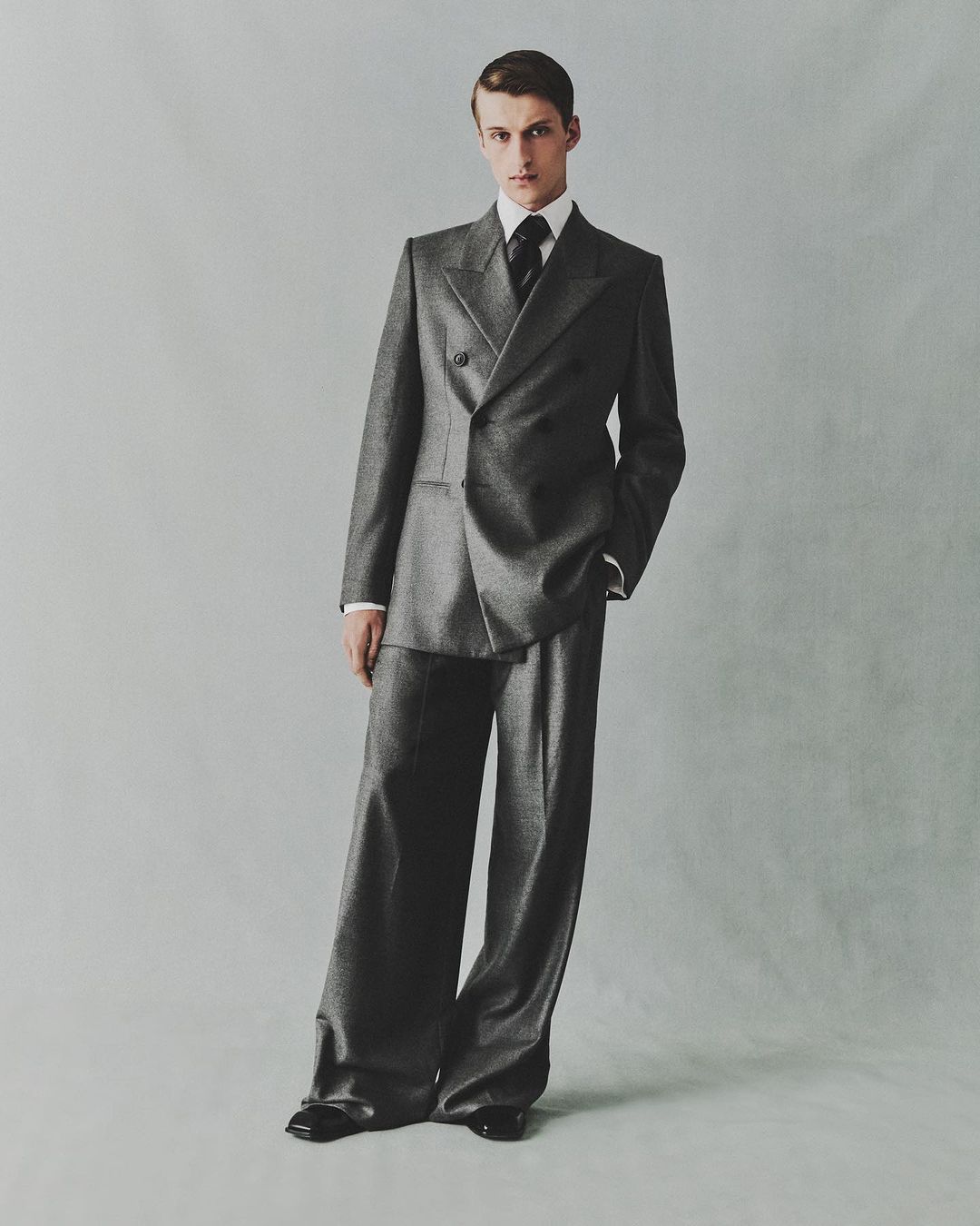 A male model poses in a grey double-breasted suit with wide-leg trousers in a studio setting against a light grey background.