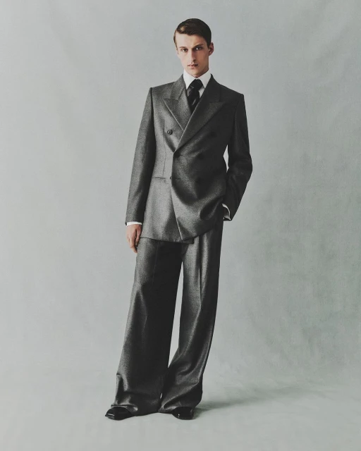 A male model poses in a grey double-breasted suit with wide-leg trousers in a studio setting against a light grey background.