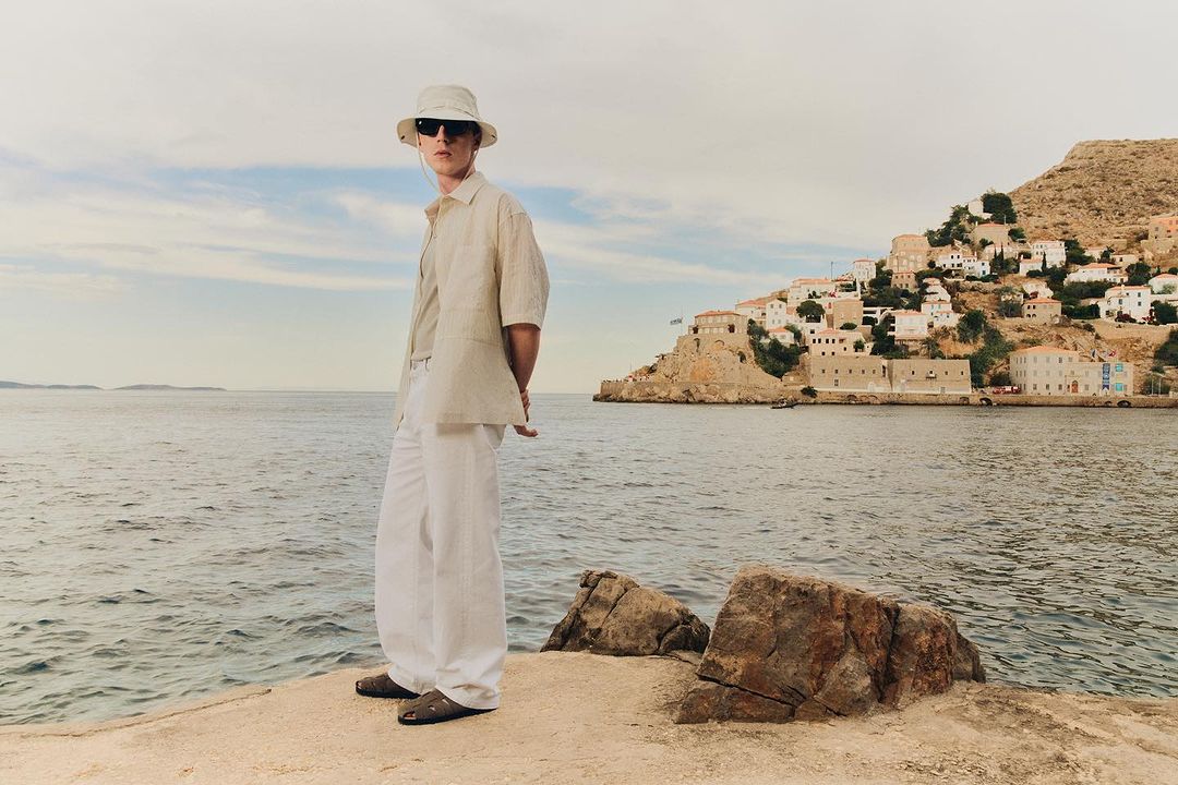 Man in linen shirt and trousers stands on a stone pier in sunglasses and bucket hat, with coastal village and water background.