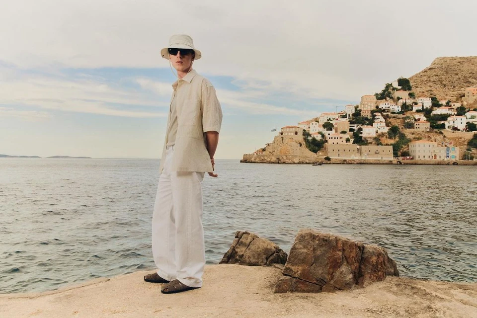Man in linen shirt and trousers stands on a stone pier in sunglasses and bucket hat, with coastal village and water background.