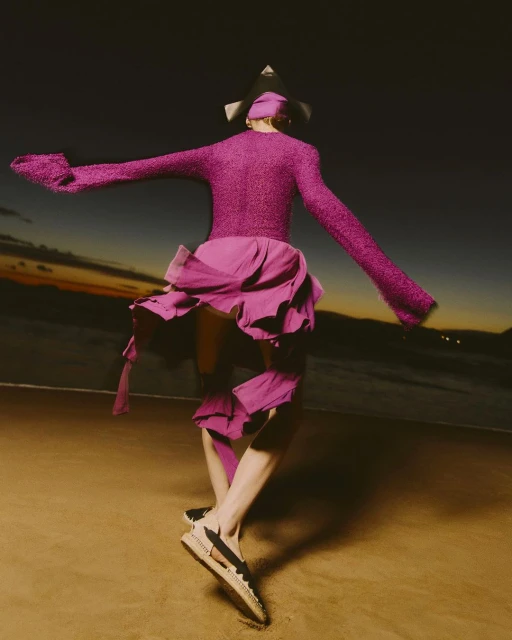 Person in magenta sweater and skirt on the beach at dusk, balancing on one foot with arms outstretched, wearing a unique geometrical hat.