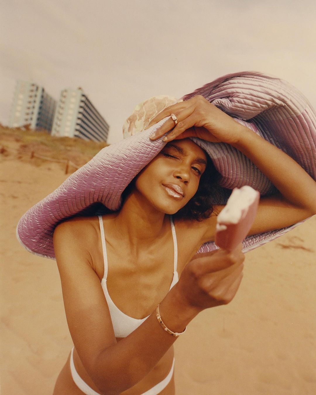 A fashionable woman on a beach under a bright sky, holding an ice cream. She wears a sun hat and a white bathing suit.