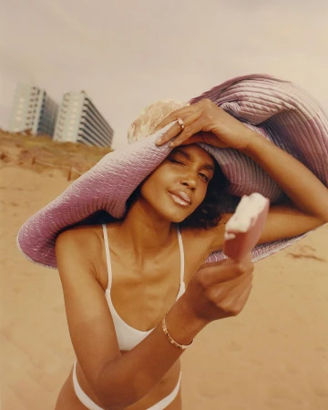 A fashionable woman on a beach under a bright sky, holding an ice cream. She wears a sun hat and a white bathing suit.