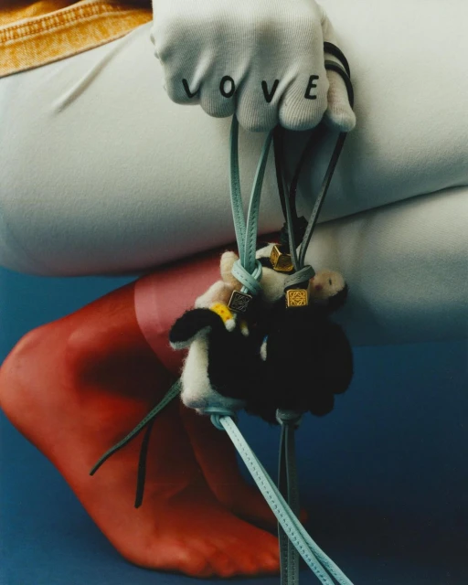 Close-up shot of a hand in a LOVE glove holding a furry plush charm attached to leather strings, with a leg and foot in the background.