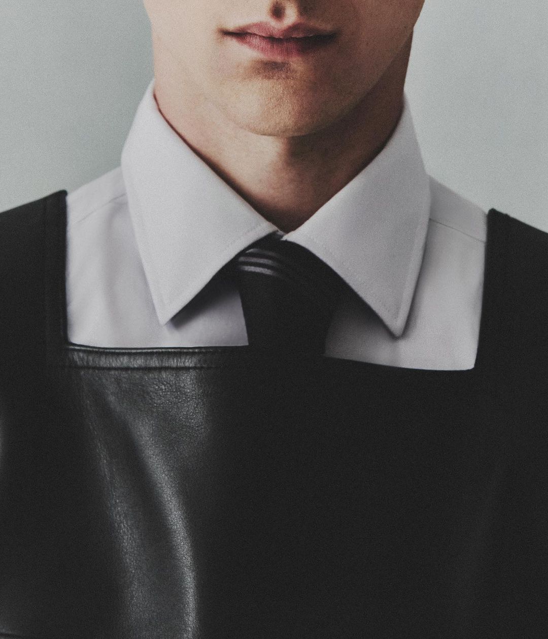 Close-up of a man wearing a black leather harness over a white shirt and a black tie, with a light gray background.
