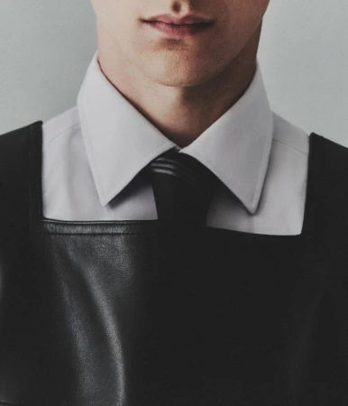 Close-up of a man wearing a black leather harness over a white shirt and a black tie, with a light gray background.