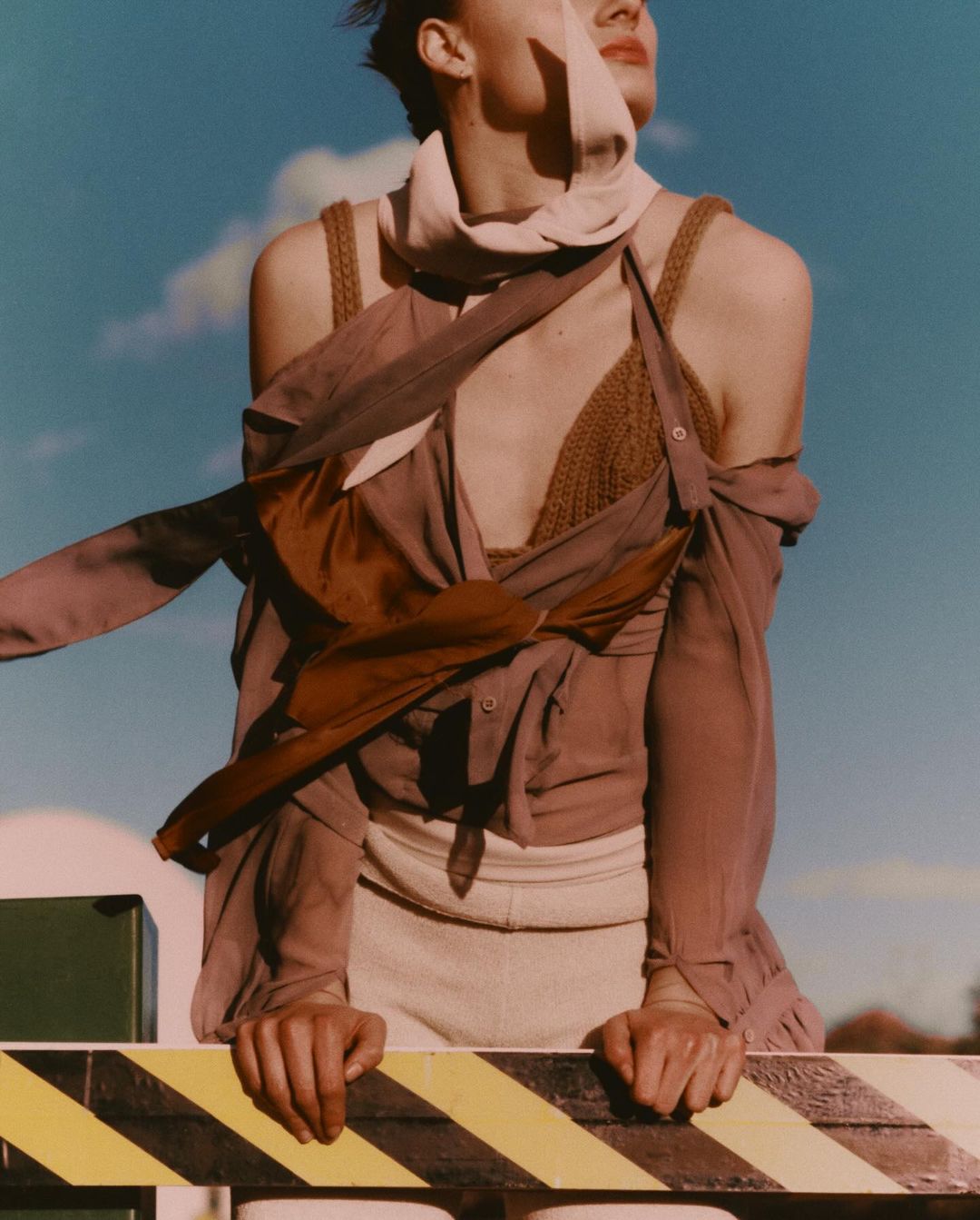 A fair-skinned model wearing a knit top and flowing shirt leans on a guardrail against a cloudy blue sky in an outdoor fashion shot.
