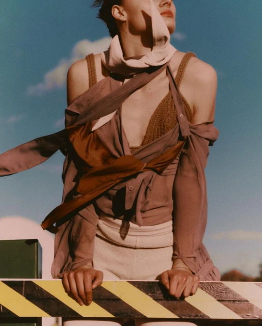 A fair-skinned model wearing a knit top and flowing shirt leans on a guardrail against a cloudy blue sky in an outdoor fashion shot.