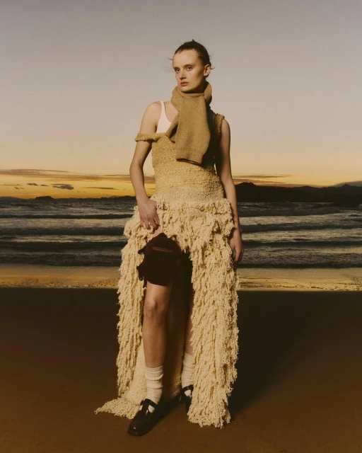 A fair-skinned woman stands on a beach at sunset wearing a tan, woven dress and scarf, paired with white socks and black shoes.