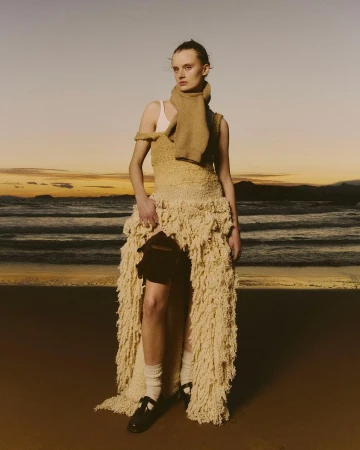 A fair-skinned woman stands on a beach at sunset wearing a tan, woven dress and scarf, paired with white socks and black shoes.