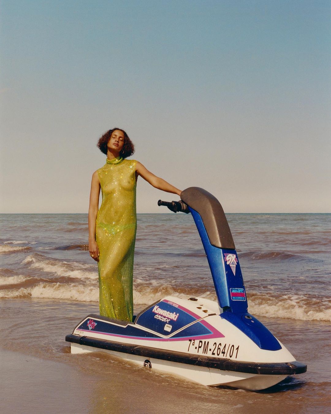 Woman in a sparkly green gown poses next to a Kawasaki jet ski on a sandy beach with a horizon line in the background under a blue sky.
