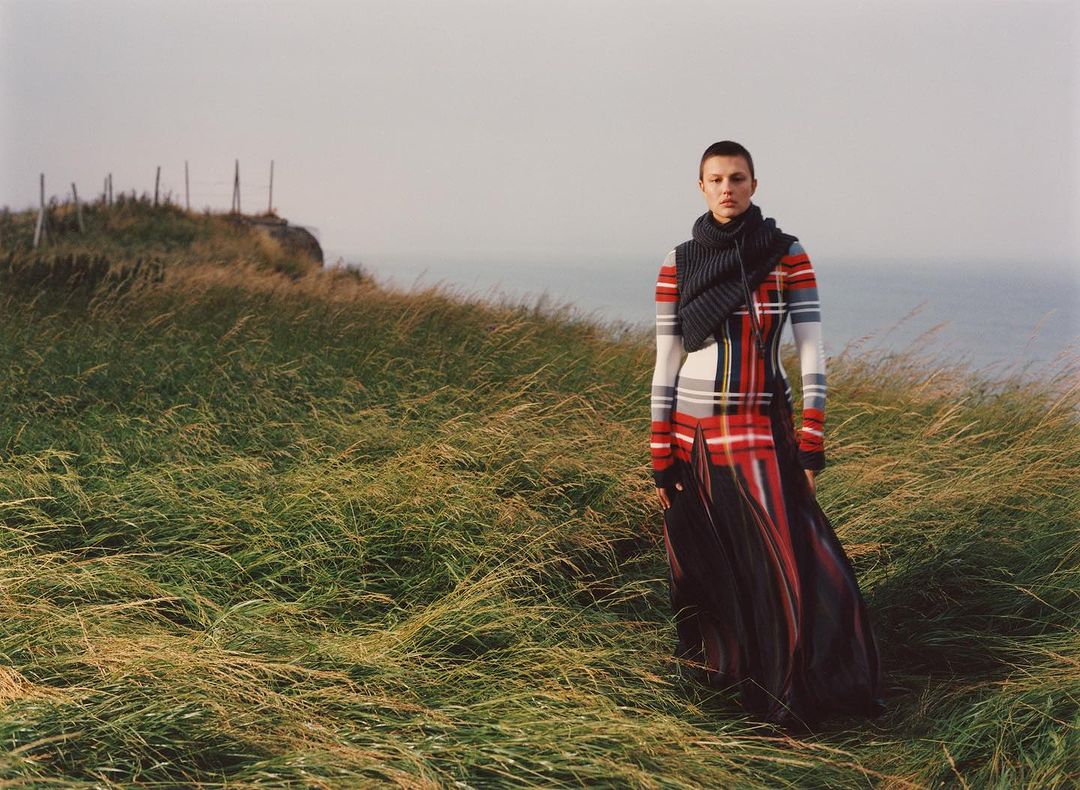 A solitary fashion model stands in a field of tall grass by the sea wearing a patterned dress and a knitted scarf on an overcast day.