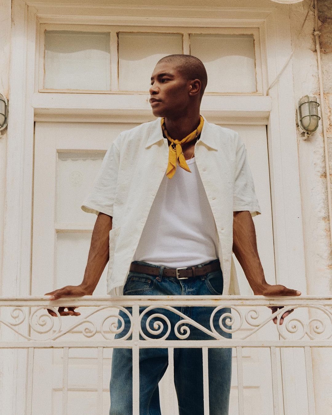 A dark-skinned male model leans against a wrought-iron balcony wearing a white shirt, yellow kerchief, and blue jeans with natural lighting.