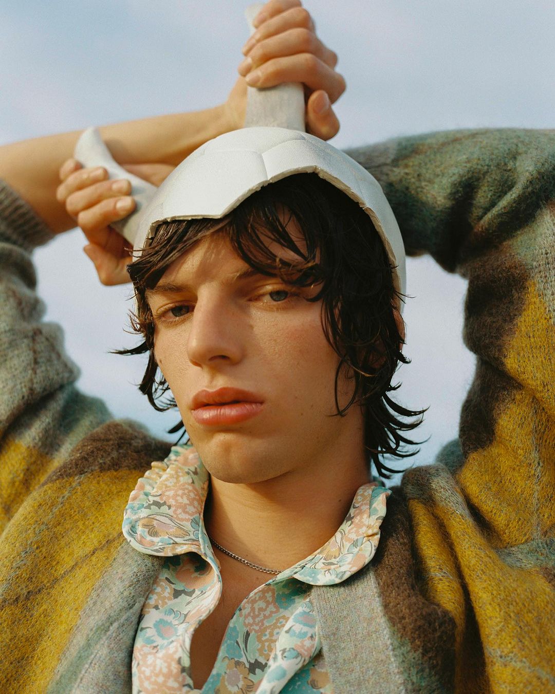Portrait of a young adult male wearing floral shirt and a gray, yellow and brown cardigan with a soccer ball helmet in an outdoor setting.