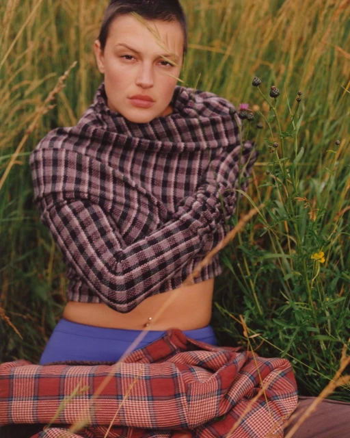Woman in plaid top and blue bottoms sits in a grassy field. She is surrounded by tall green grass and some wildflowers.