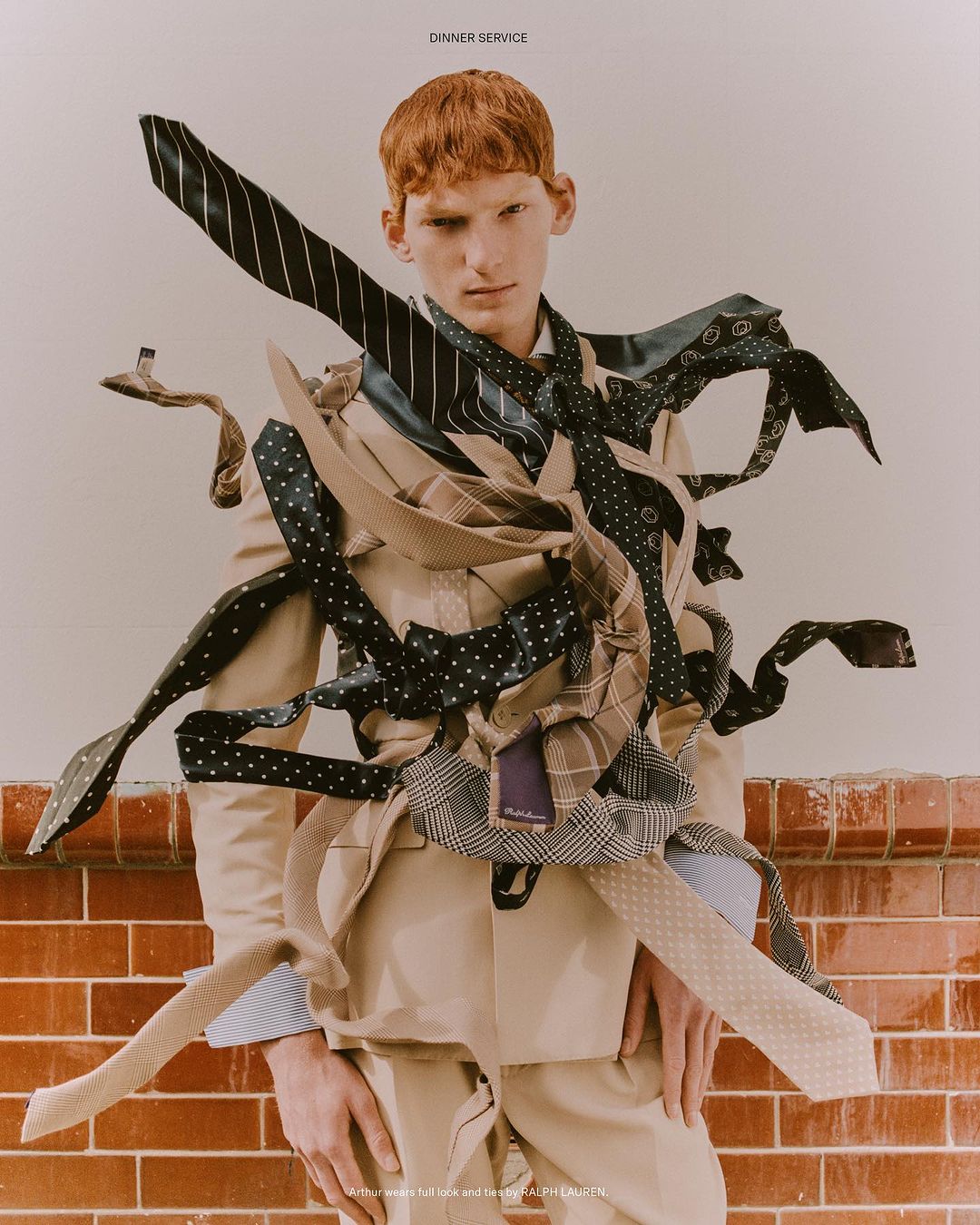 Red-haired male model in a suit jacket poses with a collection of neckties draped around him in a fashion forward studio shot.