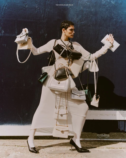 A woman in a long dress poses with various handbags attached to her body against a dark blue wall, fashion-forward, unique style.