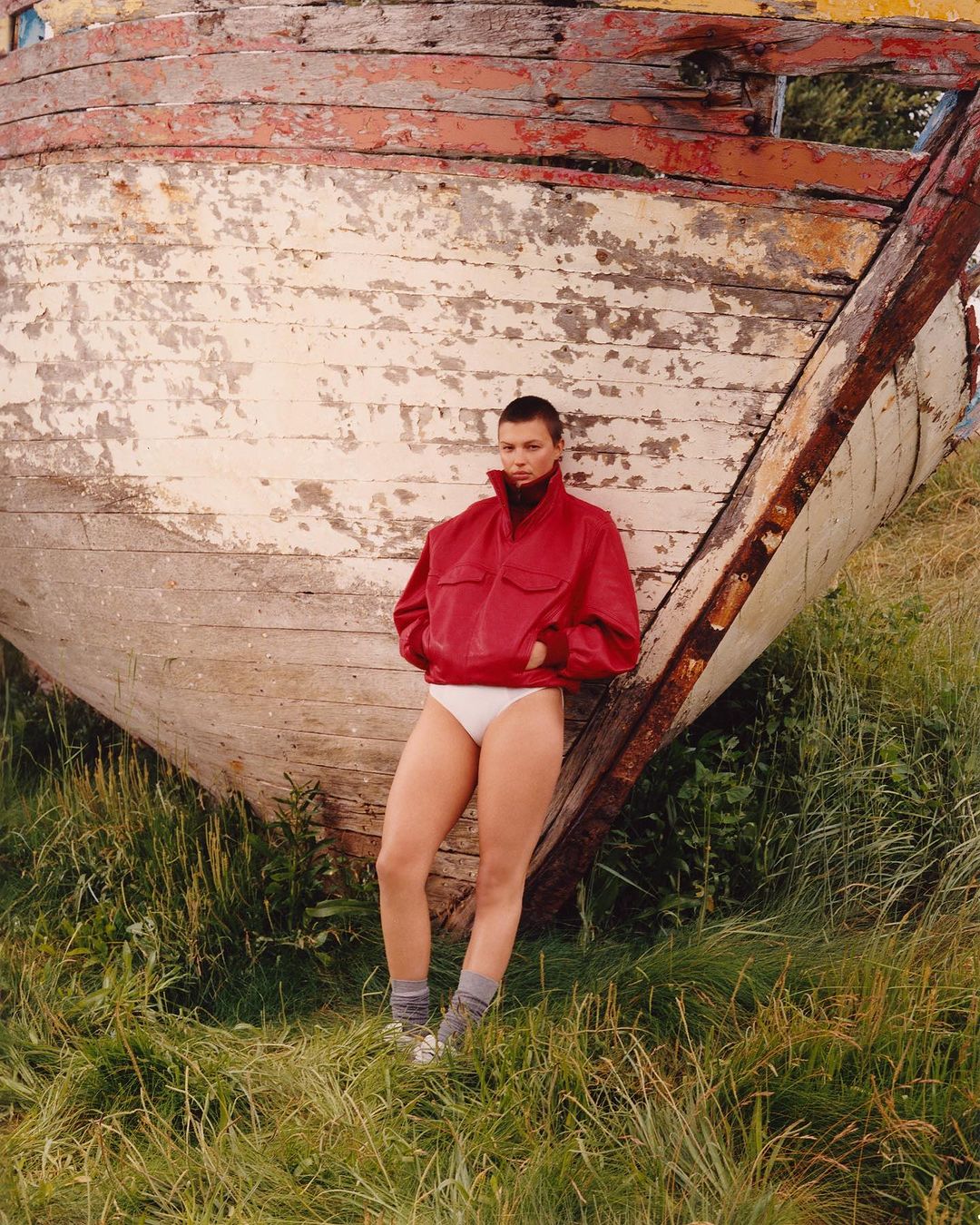 Model with shaved head wearing red jacket and white briefs leaning against weathered wooden boat in grass