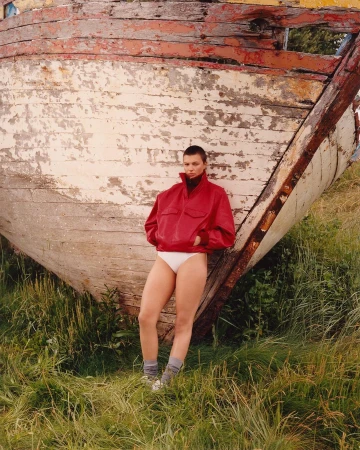 Model with shaved head wearing red jacket and white briefs leaning against weathered wooden boat in grass
