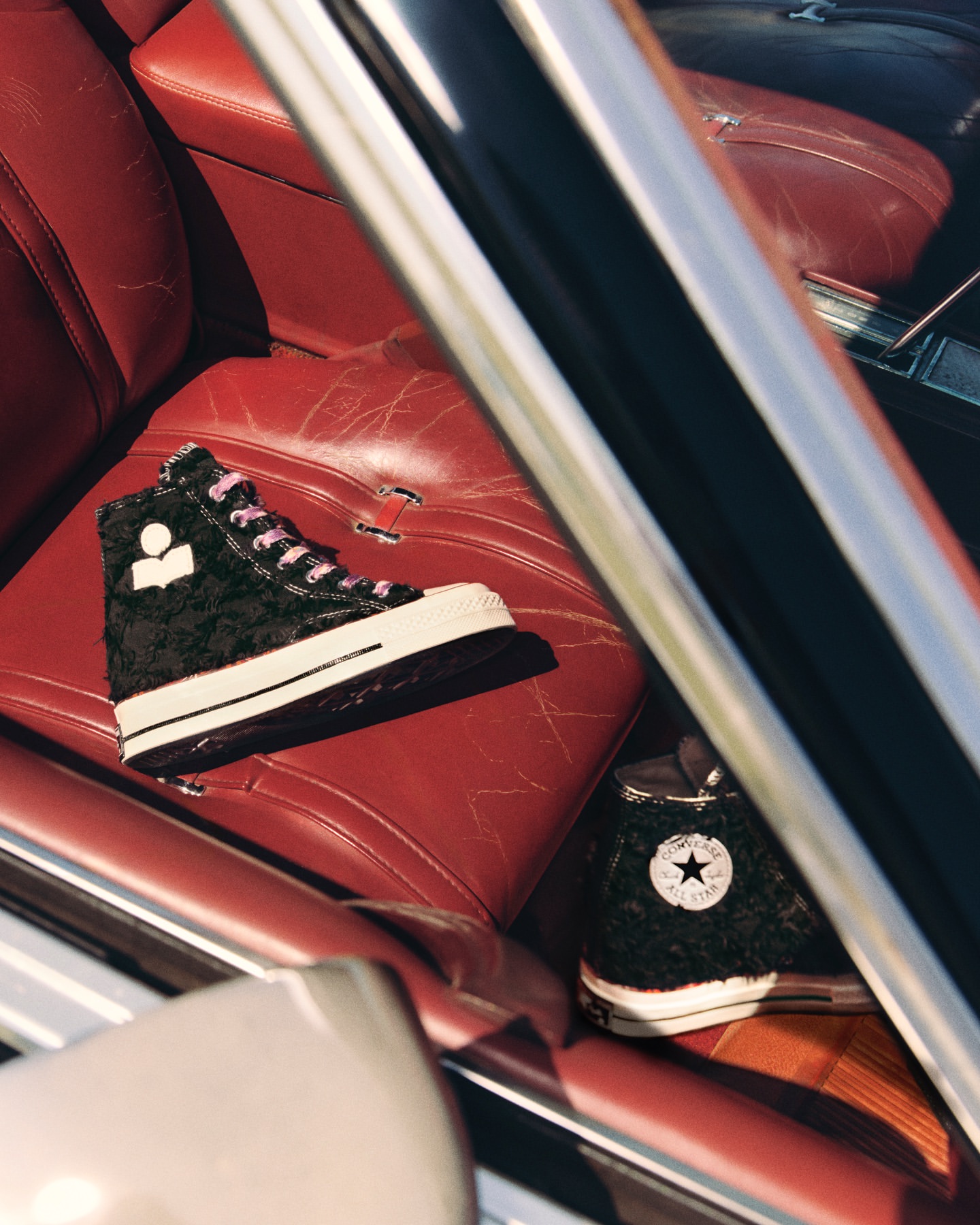 A pair of Converse high-top sneakers with fuzzy black uppers are positioned on the red leather seat inside of a car interior.