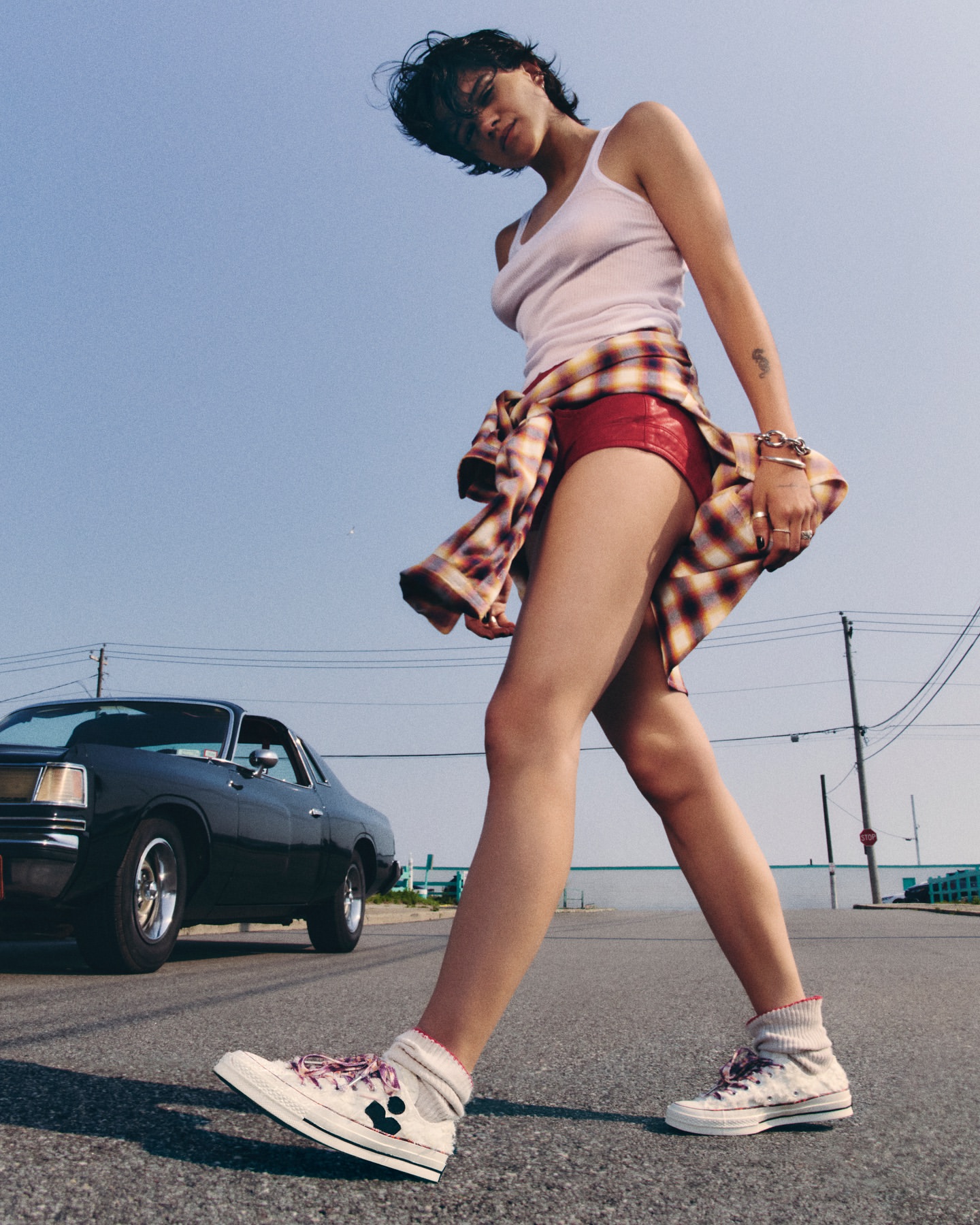 A woman in casual clothing walks on a street with a classic car in the background, under a clear blue sky, showcasing a relaxed vibe.