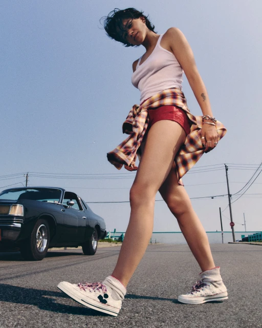 A woman in casual clothing walks on a street with a classic car in the background, under a clear blue sky, showcasing a relaxed vibe.