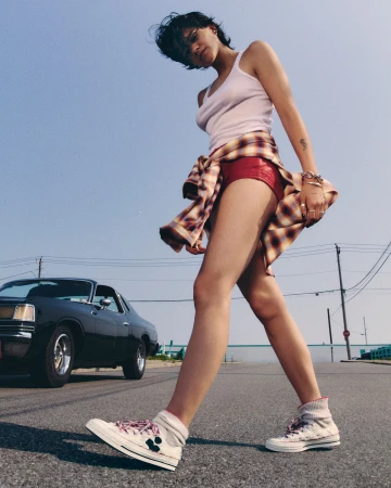 A woman in casual clothing walks on a street with a classic car in the background, under a clear blue sky, showcasing a relaxed vibe.