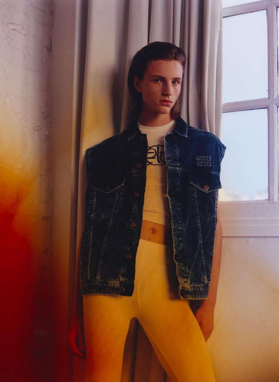 Young adult poses in front of a window wearing a white tee shirt, sleeveless denim jacket, and yellow bottoms in a studio setting.