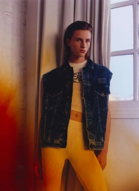 Young adult poses in front of a window wearing a white tee shirt, sleeveless denim jacket, and yellow bottoms in a studio setting.